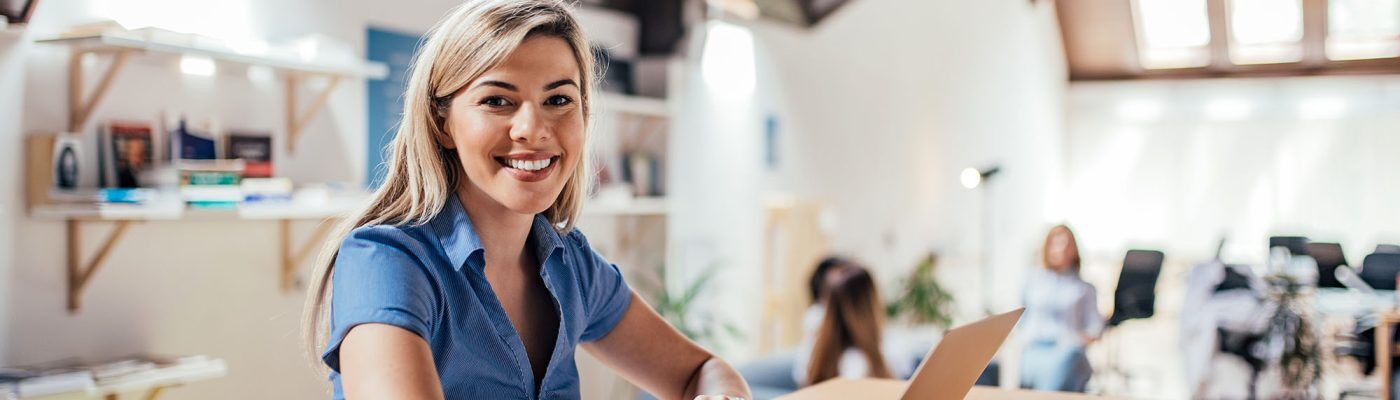 Cheerful young woman using laptop in modern environment. Looking at camera.